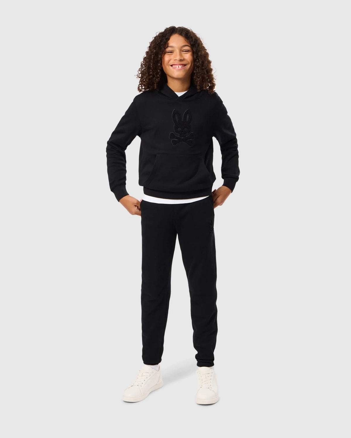 A child with curly hair smiles confidently, standing with hands on hips. They wear a black Psycho Bunny KIDS PACIFIC CHENILLE HOODIE - B0H930D200 adorned with a charming cartoon graphic, paired with black pants and white sneakers. The plain gray background accentuates the youthful energy radiating from their playful pose.