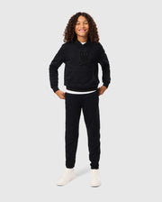 A child with curly hair smiles confidently, standing with hands on hips. They wear a black Psycho Bunny KIDS PACIFIC CHENILLE HOODIE - B0H930D200 adorned with a charming cartoon graphic, paired with black pants and white sneakers. The plain gray background accentuates the youthful energy radiating from their playful pose.