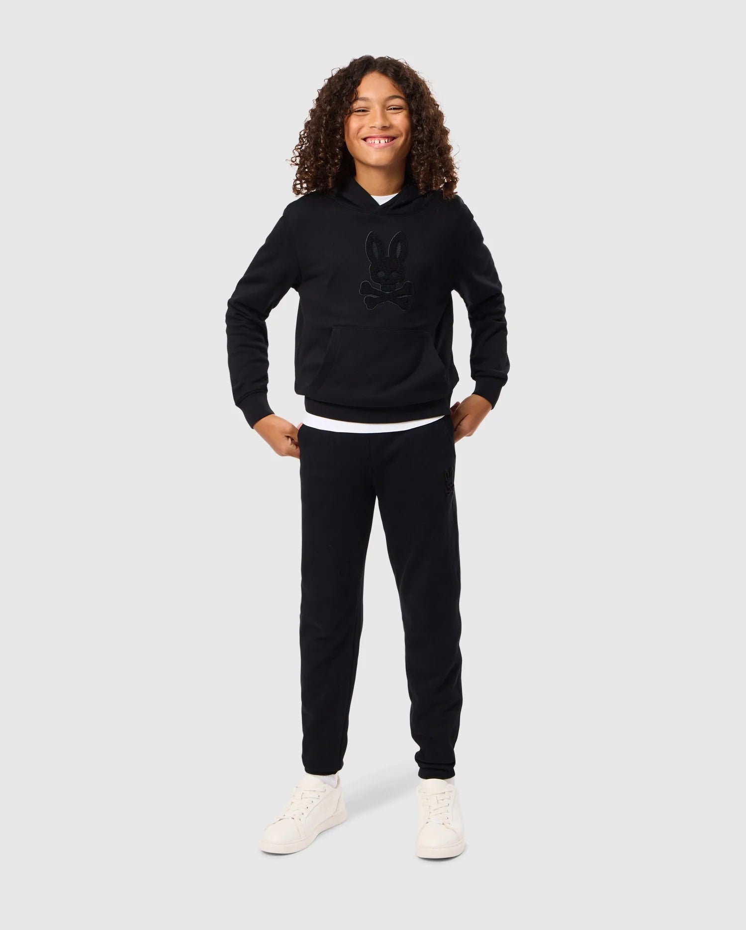 A child with curly hair smiles confidently, standing with hands on hips. They wear a black Psycho Bunny KIDS PACIFIC CHENILLE HOODIE - B0H930D200 adorned with a charming cartoon graphic, paired with black pants and white sneakers. The plain gray background accentuates the youthful energy radiating from their playful pose.