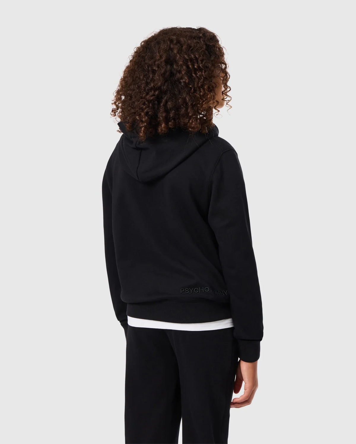 A person with curly hair, wearing a black KIDS PACIFIC CHENILLE HOODIE - B0H930D200 by Psycho Bunny and black pants, stands facing away from the camera against a gray background.