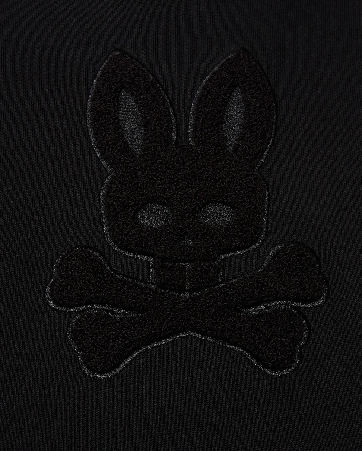 The KIDS PACIFIC CHENILLE HOODIE - B0H930D200 by Psycho Bunny highlights a black-on-black embroidered design, featuring the outline of a rabbit head above crossbones against a dark fabric background.
