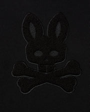 The KIDS PACIFIC CHENILLE HOODIE - B0H930D200 by Psycho Bunny highlights a black-on-black embroidered design, featuring the outline of a rabbit head above crossbones against a dark fabric background.