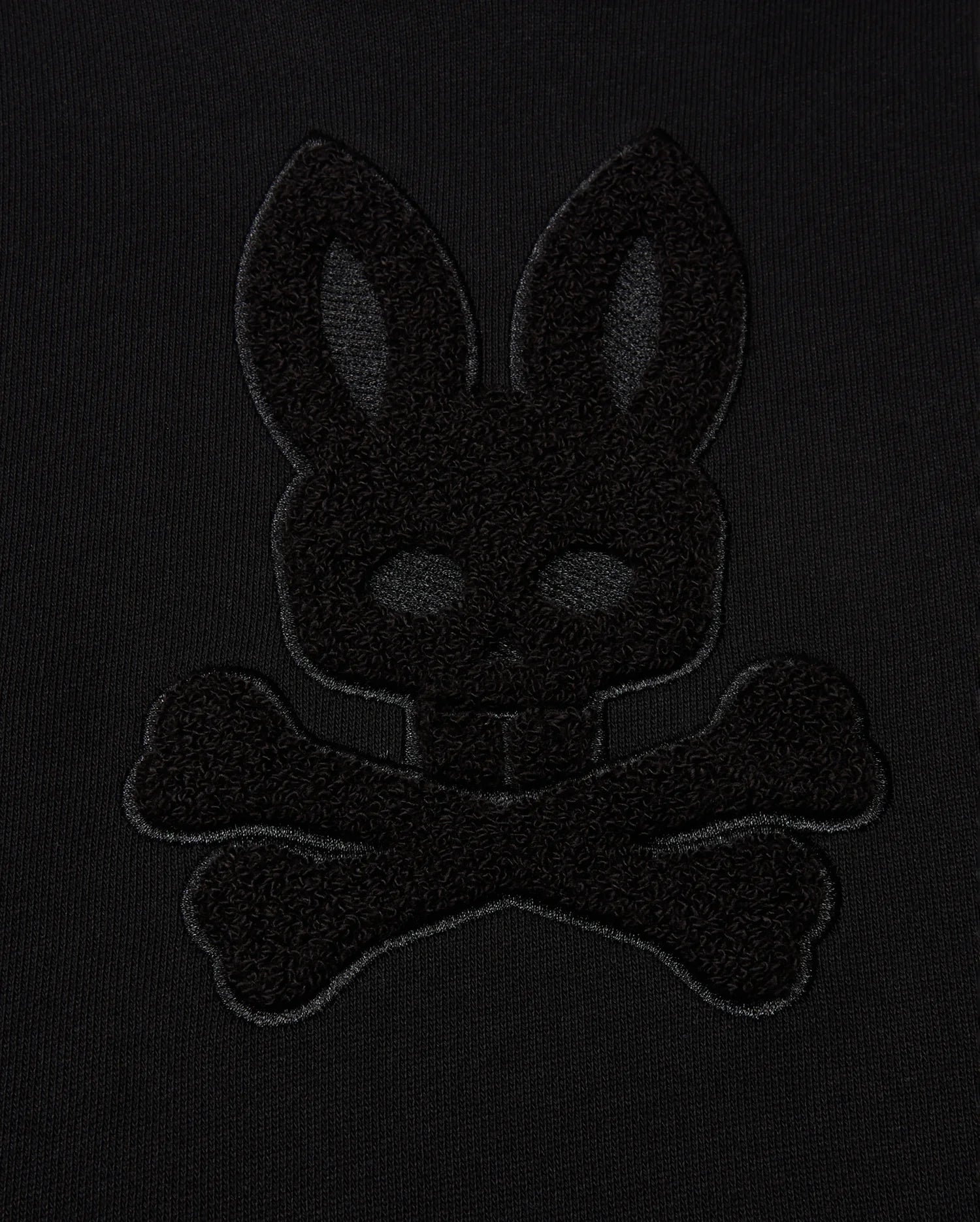The KIDS PACIFIC CHENILLE HOODIE - B0H930D200 by Psycho Bunny highlights a black-on-black embroidered design, featuring the outline of a rabbit head above crossbones against a dark fabric background.
