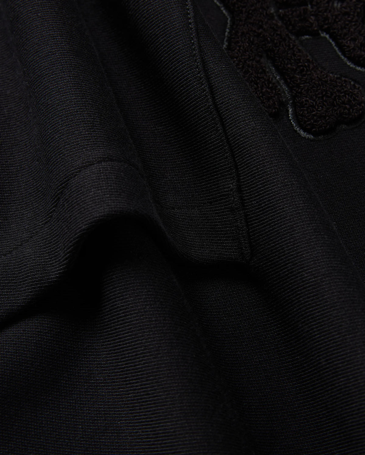 Here's a close-up of the black textured fabric from the KIDS PACIFIC CHENILLE HOODIE by Psycho Bunny (B0H930D200). The visible seam and part of the raised chenille embroidery design stand out. The soft fabric is slightly folded, emphasizing stitching details that evoke the cozy feel of a French terry hoodie.