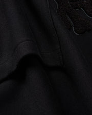 Here's a close-up of the black textured fabric from the KIDS PACIFIC CHENILLE HOODIE by Psycho Bunny (B0H930D200). The visible seam and part of the raised chenille embroidery design stand out. The soft fabric is slightly folded, emphasizing stitching details that evoke the cozy feel of a French terry hoodie.