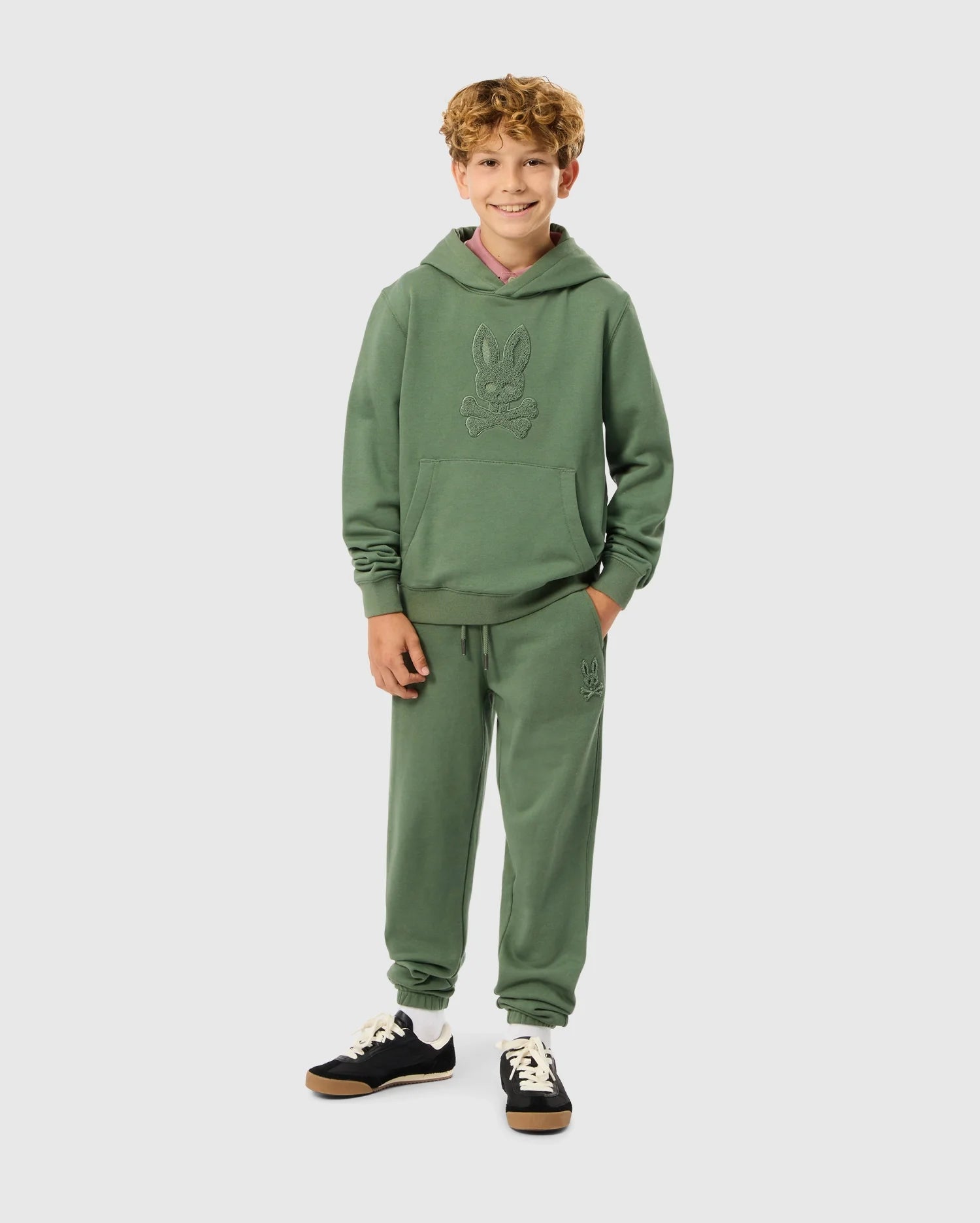 A young person with curly hair stands smiling, wearing the KIDS PACIFIC CHENILLE HOODIE (B0H930D200) and matching joggers from Psycho Bunny. The outfit features a subtle rabbit design, and they complete their stylish look with black sneakers featuring white laces.