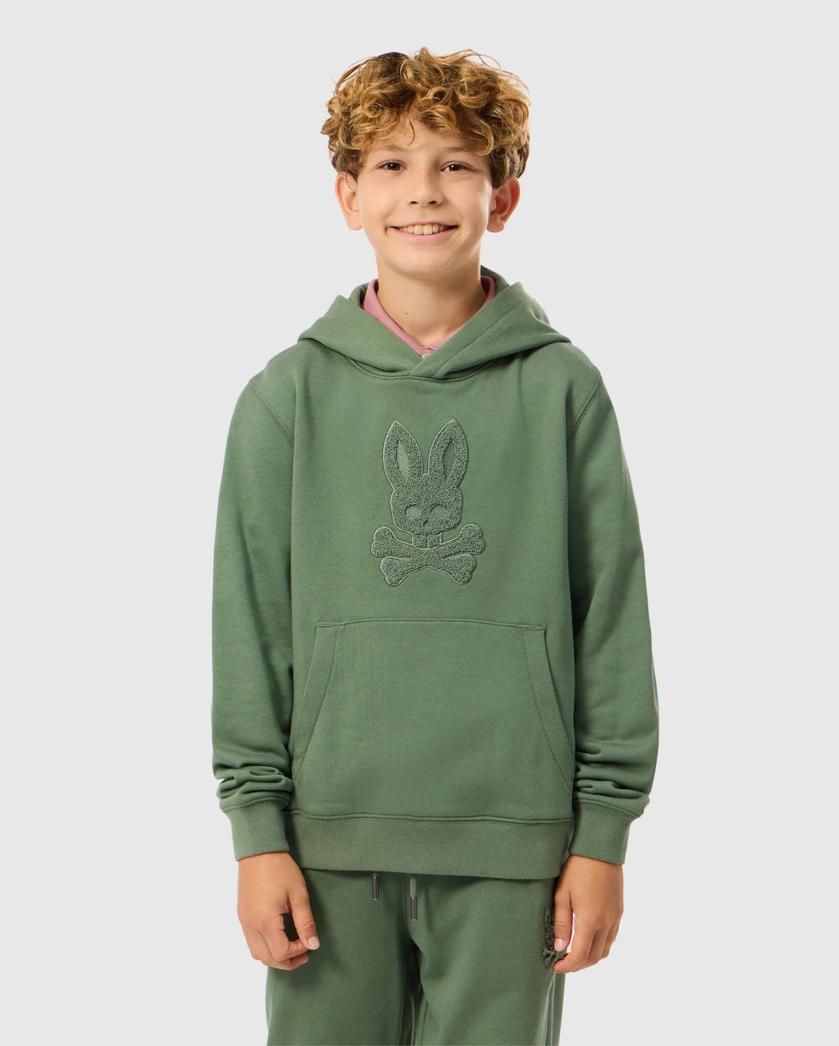 A smiling child with curly hair is wearing the KIDS PACIFIC CHENILLE HOODIE - B0H930D200, by Psycho Bunny. The green French terry hoodie showcases a distinctive design with chenille embroidery. Standing against a plain background, the child has their hands in the front pockets, giving off a casual and comfortable vibe.