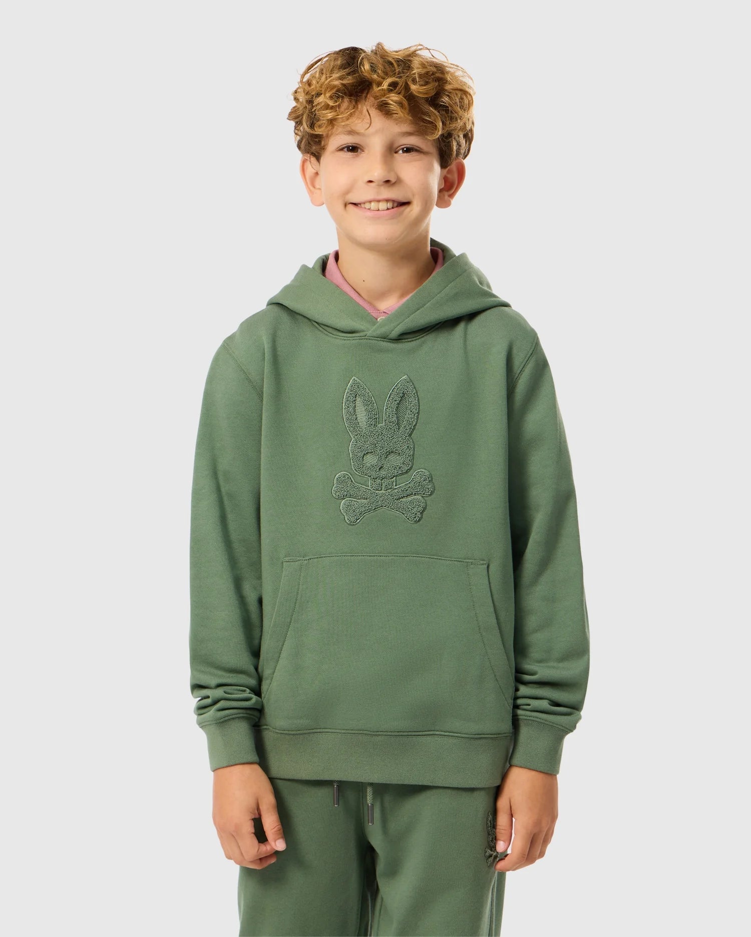 A smiling child with curly hair is wearing the KIDS PACIFIC CHENILLE HOODIE - B0H930D200, by Psycho Bunny. The green French terry hoodie showcases a distinctive design with chenille embroidery. Standing against a plain background, the child has their hands in the front pockets, giving off a casual and comfortable vibe.