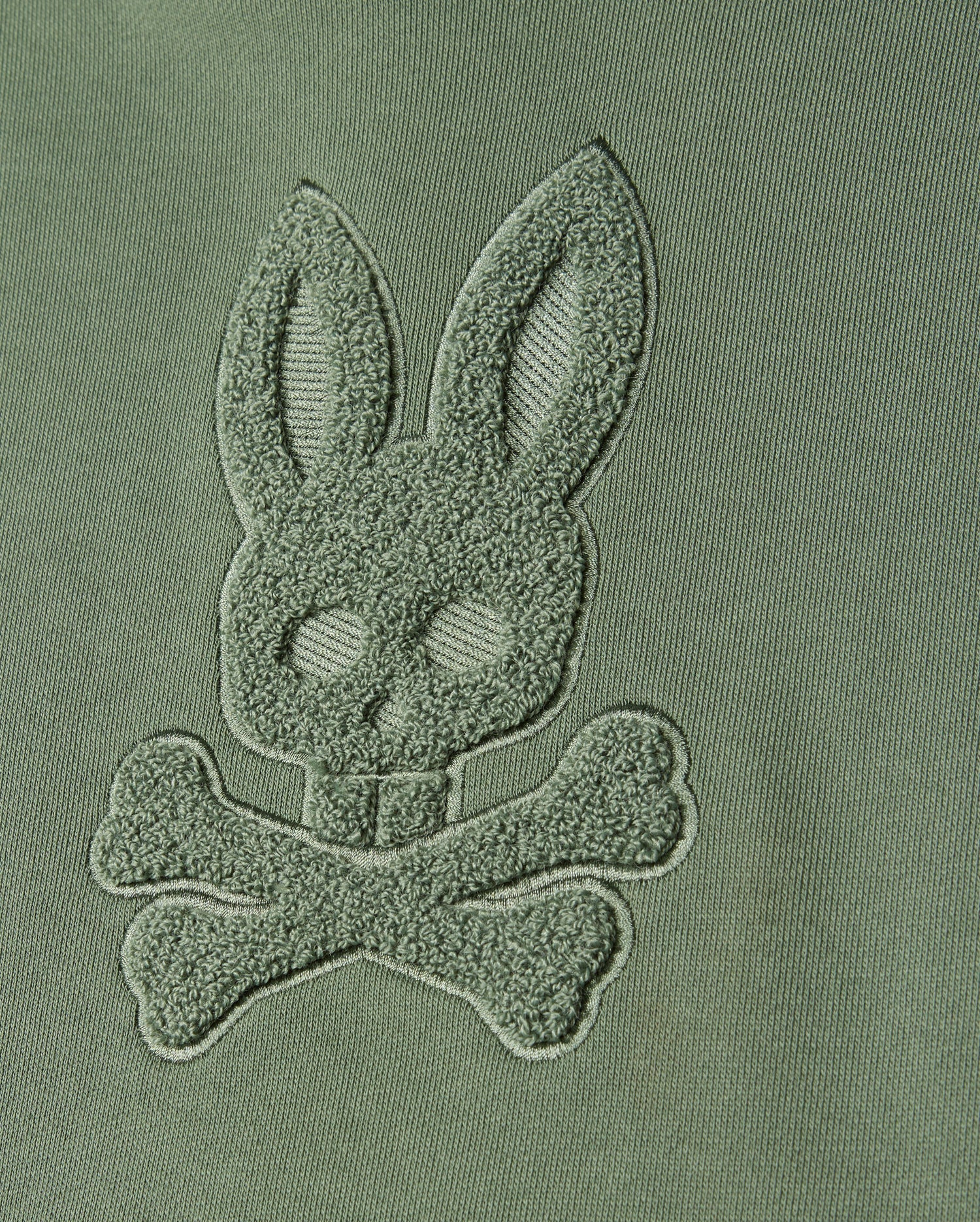Featuring a close-up of green fabric adorned with chenille embroidery, the design presents a bunny head and crossed bones. This motif captures the essence of Psycho Bunny's distinct style, seamlessly blending into the fabric for a textured, raised monochromatic appearance—ideal for creating a masterpiece like the KIDS PACIFIC CHENILLE HOODIE (B0H930D200) from Psycho Bunny.