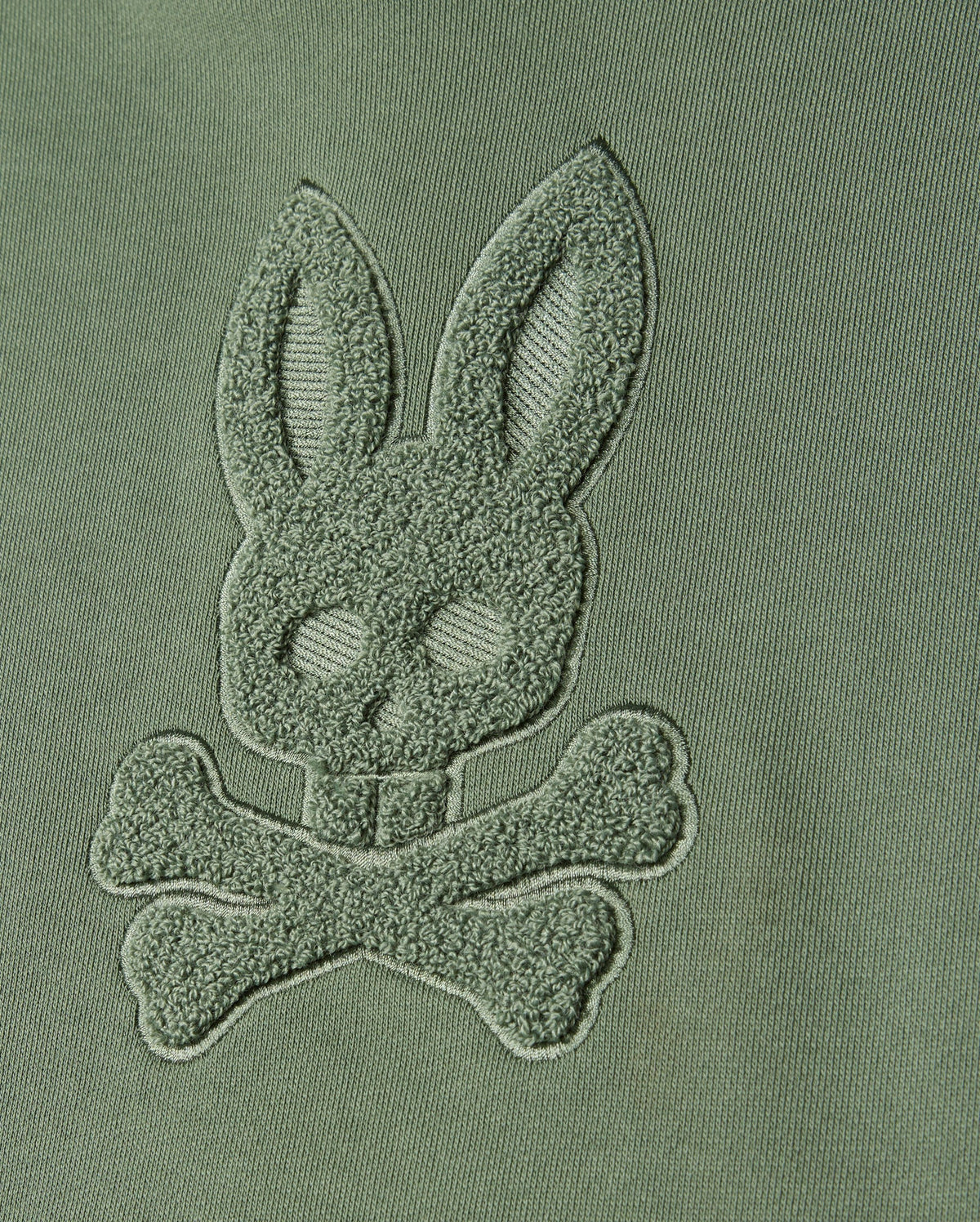 Featuring a close-up of green fabric adorned with chenille embroidery, the design presents a bunny head and crossed bones. This motif captures the essence of Psycho Bunny's distinct style, seamlessly blending into the fabric for a textured, raised monochromatic appearance—ideal for creating a masterpiece like the KIDS PACIFIC CHENILLE HOODIE (B0H930D200) from Psycho Bunny.
