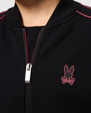 Close-up of a person wearing the KIDS TRAVIS TRACK JACKET - B0J142D200 by Psycho Bunny. The black regular fit jacket boasts a red stitched bunny logo and crossbones on the left side of the chest. Made from high-quality cotton-blend, it features a silver zipper with an engraved bunny emblem. Red stitching details accentuate the shoulders.