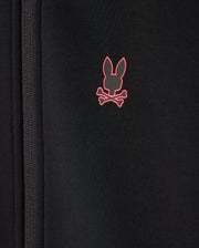 A close-up of the KIDS TRAVIS TRACK JACKET - B0J142D200 by Psycho Bunny, designed in a regular fit and crafted from high-quality cotton-blend fabric with a zipper on the left side. It features a small pink logo of a rabbit's head with crossbones underneath on the right side.