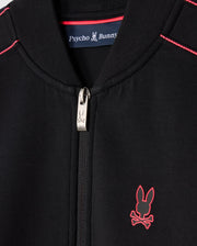 Close-up of a black Kids Travis Track Jacket (B0J142D200) by Psycho Bunny, featuring red stitching and a small red emblem of a bunny with crossbones. Made from high-quality cotton-blend, it includes a zipper pull adorned with the same bunny design. Inside the collar, a label reads "Psycho Bunny," ensuring its regular fit.