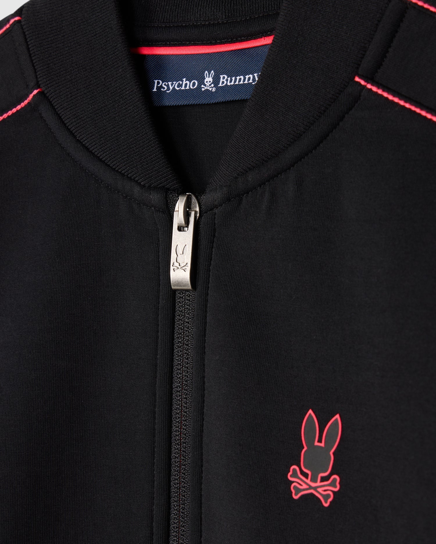 Close-up of a black Kids Travis Track Jacket (B0J142D200) by Psycho Bunny, featuring red stitching and a small red emblem of a bunny with crossbones. Made from high-quality cotton-blend, it includes a zipper pull adorned with the same bunny design. Inside the collar, a label reads "Psycho Bunny," ensuring its regular fit.