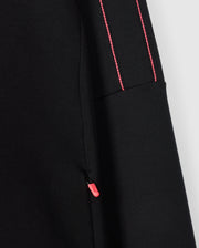 Close-up of the Psycho Bunny Kids Travis Track Jacket (B0J142D200), crafted from a high-quality cotton-blend fabric. The jacket features a small pink zipper pull on the pocket seam, and the sleeve showcases two thin, parallel pink stitches running vertically, adding a contrasting detail to the predominantly black fabric. The texture appears smooth and slightly stretchy.