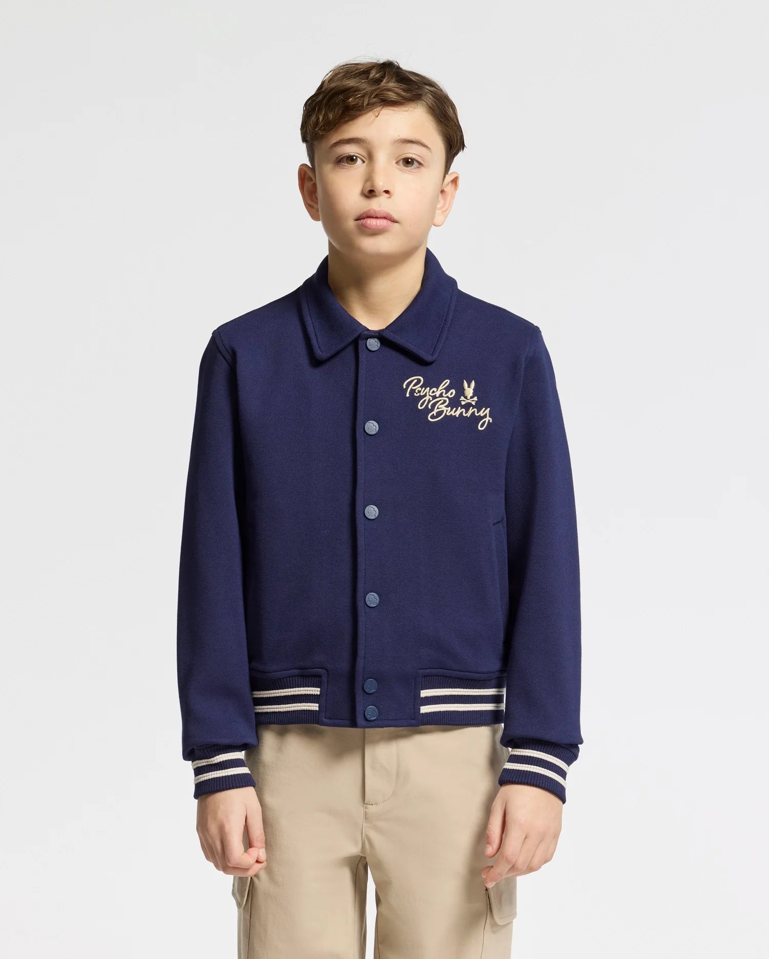 A young person wears the Psycho Bunny Kids Desert Coach Bomber Jacket (B0J428F200) in Evening Blue, featuring a collar, button-up front, embroidered chest logo, and striped cuffs and hem. They pair it with beige pants against a plain white background.
