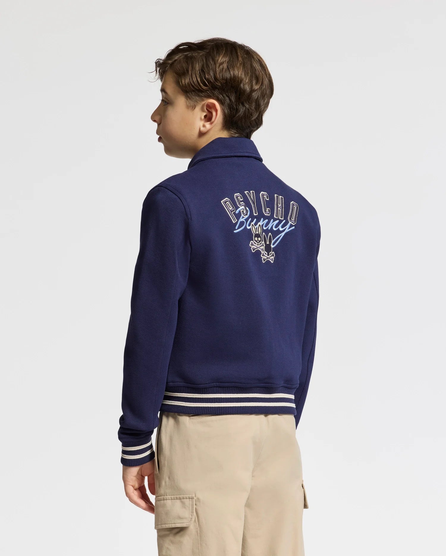 A young person with light brown hair wears the "KIDS DESERT COACH BOMBER JACKET - B0J428F200" in navy, featuring a Psycho Bunny logo on the back. It's paired with khaki pants as they stand facing away against a plain white background.