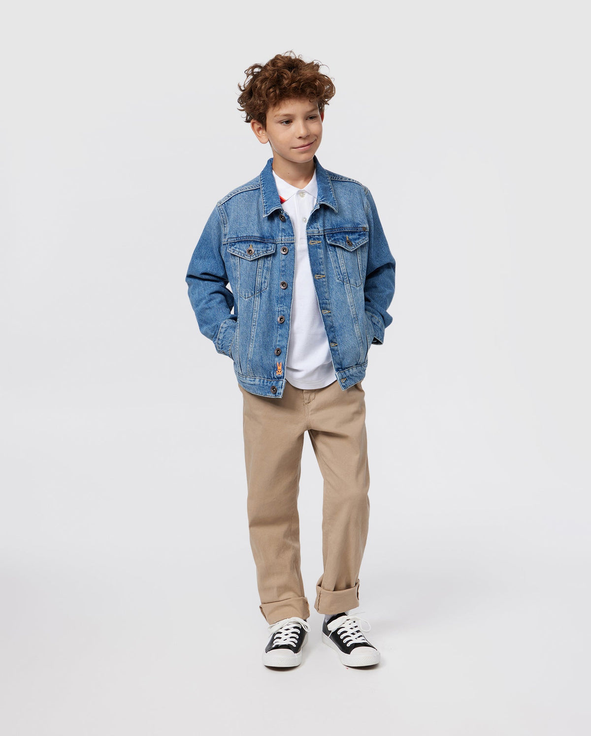 A young boy with curly hair is wearing a KIDS PATHE DENIM JACKET - B0J741X1CO by Psycho Bunny made from upcycled cotton over a white polo shirt, beige pants, and black-and-white sneakers. He stands with his hands in the jacket pockets, looking off to the side against a plain white background.