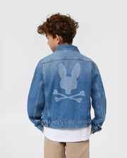 A young person with curly hair is seen from the back, wearing a blue KIDS PATHE DENIM JACKET - B0J741X1CO by Psycho Bunny. The jacket, made of upcycled cotton, features a large white logo of a bunny head with crossbones underneath. The person is also sporting khaki pants and a white shirt. The background is plain white.