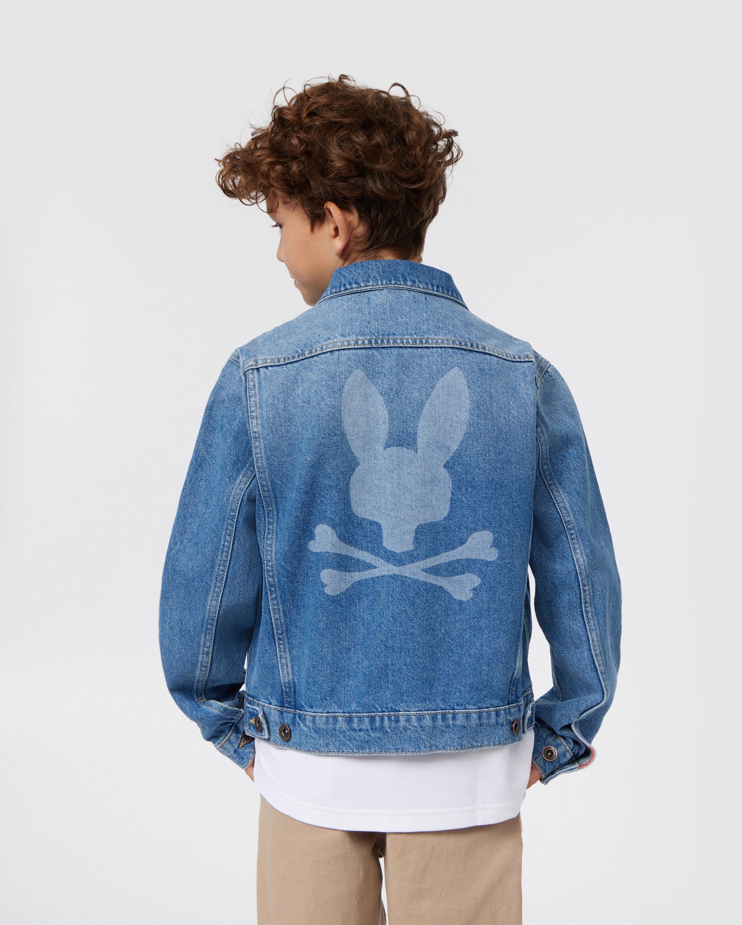 A young person with curly hair is seen from the back, wearing a blue KIDS PATHE DENIM JACKET - B0J741X1CO by Psycho Bunny. The jacket, made of upcycled cotton, features a large white logo of a bunny head with crossbones underneath. The person is also sporting khaki pants and a white shirt. The background is plain white.
