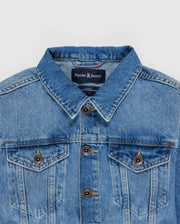 A close-up of a KIDS PATHE DENIM JACKET - B0J741X1CO made from upcycled cotton featuring a folded collar and buttoned front. The label inside reads "Psycho Bunny" with a small bunny logo above it. The jacket has chest pockets with buttoned flaps and visible stitching along the seams.