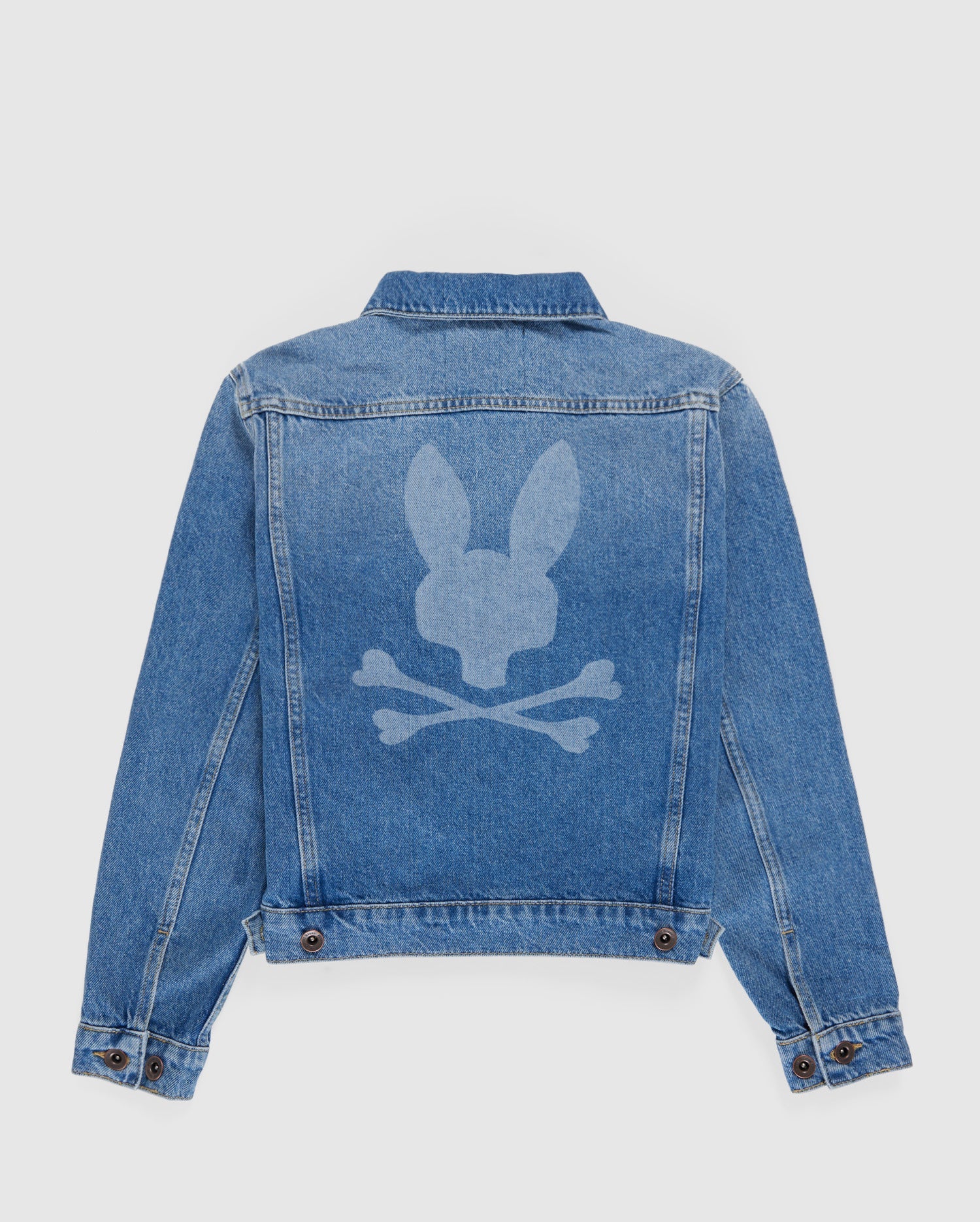 A light blue KIDS PATHE DENIM JACKET - B0J741X1CO from Psycho Bunny is shown from the back, featuring a unique design. The design, in a lighter denim wash, depicts a bunny head above two crossed bones, resembling a playful twist on the classic skull and crossbones symbol. The jacket is made from upcycled cotton for an eco-friendly touch.
