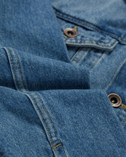 Close-up of blue denim fabric from a Psycho Bunny KIDS PATHE DENIM JACKET - B0J741X1CO, showing detailed stitching and two metal buttons. The fabric is folded, creating soft creases and highlighting the texture of the material. The image captures the classic look of upcycled cotton denim, emphasizing its weave and durability.