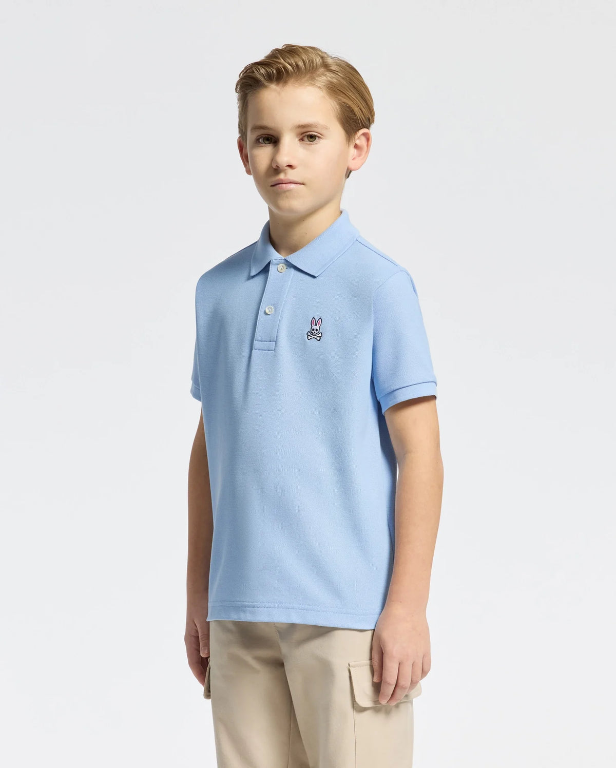 A young boy with short light brown hair stands against a white background, wearing a light blue Psycho Bunny KIDS CLASSIC PIQUE POLO (B0K001B200) with an embroidered Bunny logo on the chest, perfectly paired with khaki pants.
