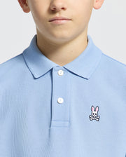 A person wearing the Psycho Bunny KIDS CLASSIC PIQUE POLO - B0K001B200 with an embroidered pink bunny and crossed bones, showcased in a cropped light blue image focusing on the lower face, neck, and upper torso.