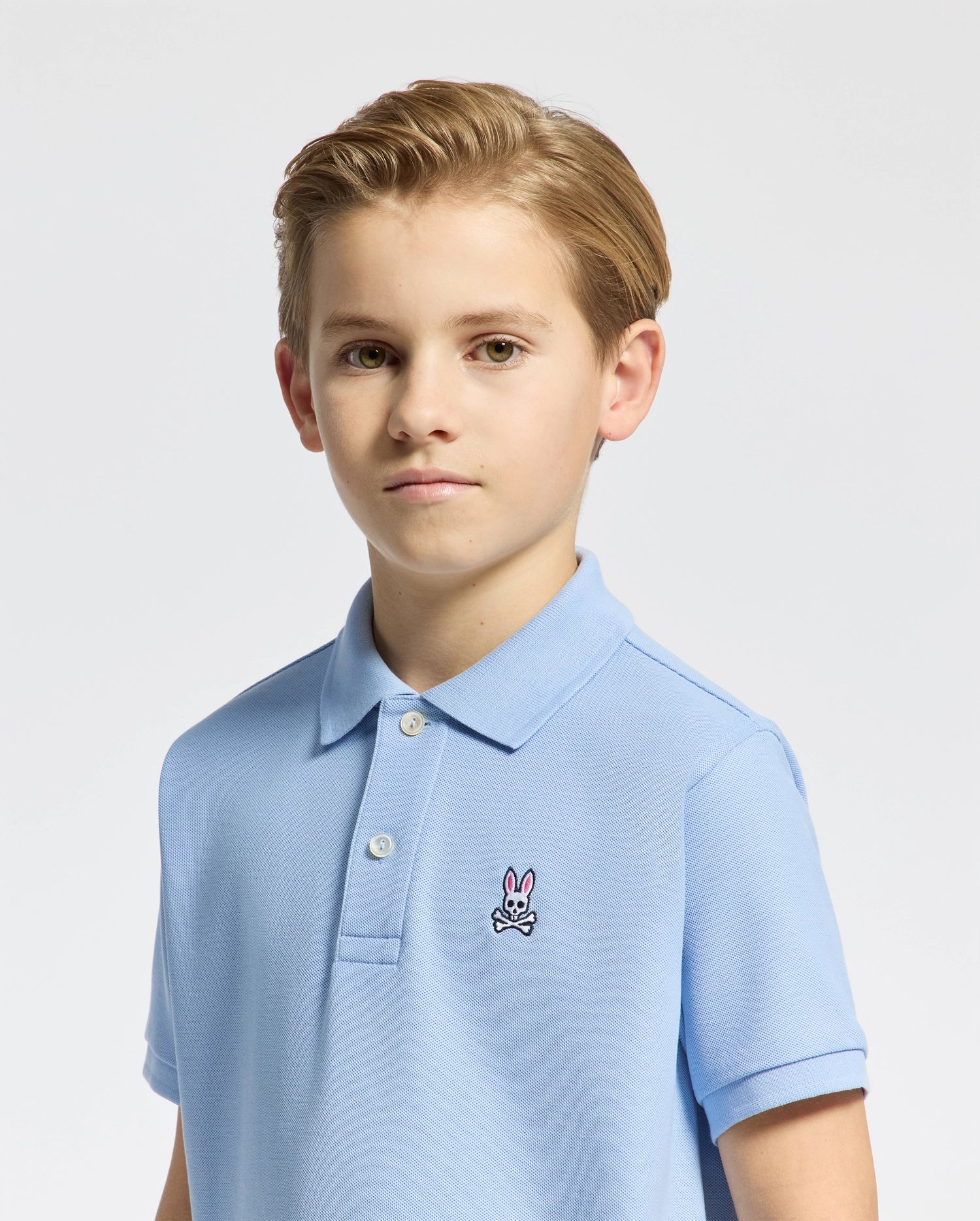 A boy with short brown hair wears a light blue KIDS CLASSIC PIQUE POLO by Psycho Bunny, adorned with a small embroidered bunny logo on the chest, set against a plain white background.