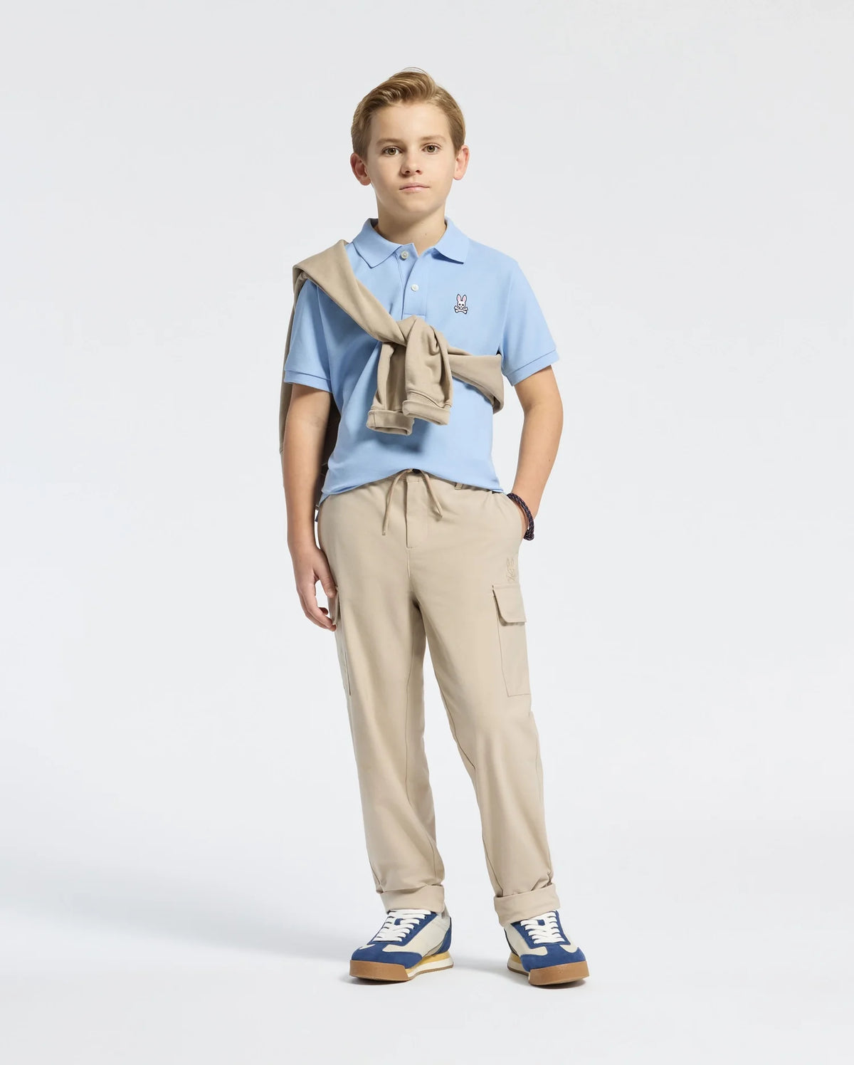 A young boy with short blond hair and a neutral expression stands against a plain background. He wears Psycho Bunny's Kids Classic Pique Polo (B0K001B200) in light blue, khaki pants, and blue sneakers. A beige sweater is draped over his shoulder, with subtle embroidered Bunny details enhancing his look.