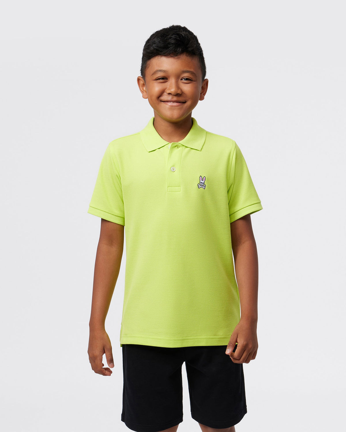 A smiling child, confident against a plain background, wears the Psycho Bunny KIDS CLASSIC POLO - B0K001CRPC in bright green. The regular fit Pima cotton piqué features a small emblem on the left side, enhancing their cheerful appearance.