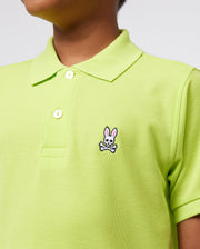 Someone is wearing a lime green Psycho Bunny Kids Classic Polo made from soft Pima cotton. It features a small pink and white bunny logo above crossed bones on the chest, has buttons, a collar, and a regular fit in front of a plain background.