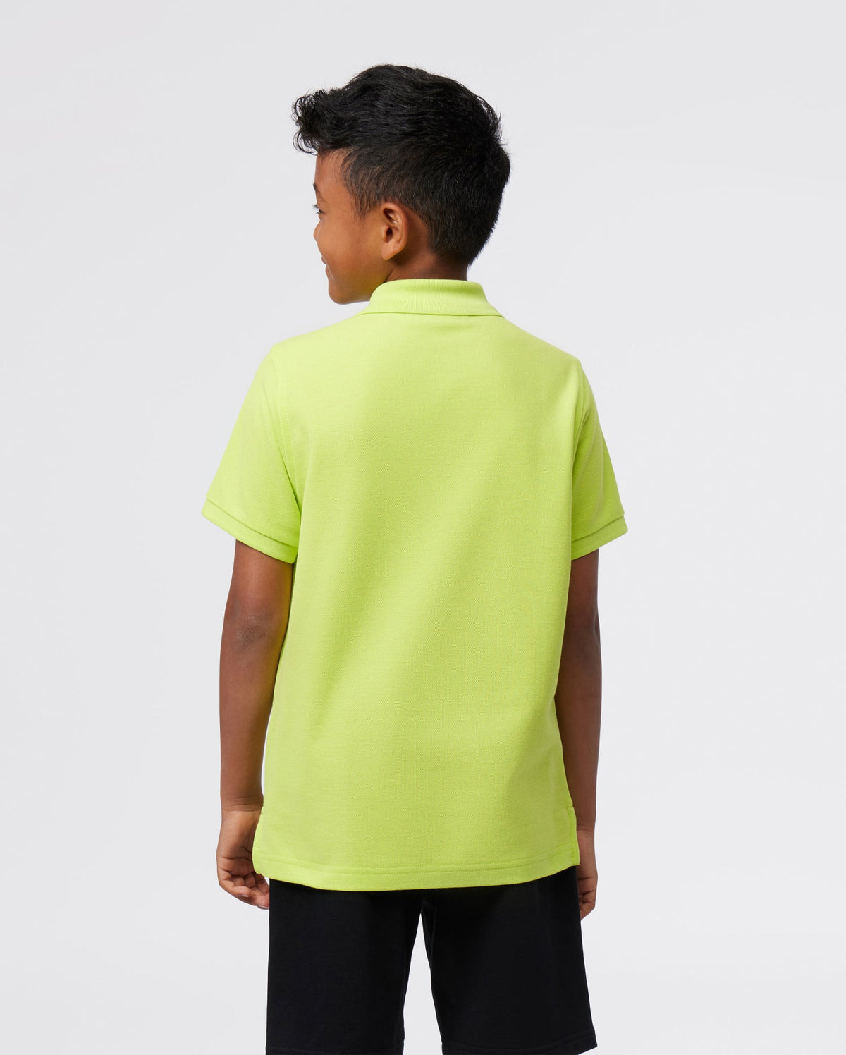A young boy stands with his back to the camera, wearing a Psycho Bunny KIDS CLASSIC POLO - B0K001CRPC in light green made from soft Pima cotton, paired with black shorts against a plain white background.