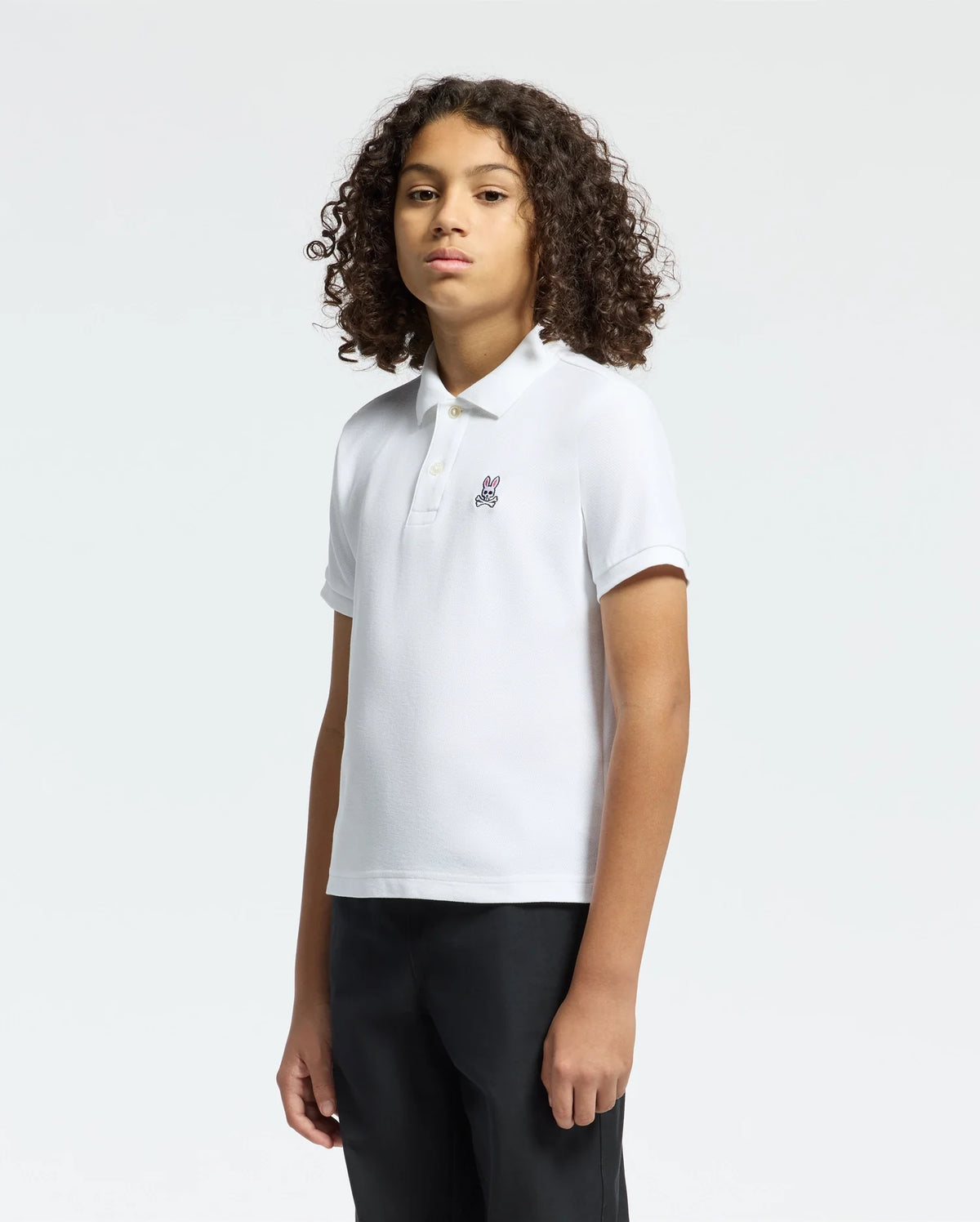 A curly-haired person models Psycho Bunny's KIDS CLASSIC PIQUE POLO SHIRT - B0K001CRPC against a white background, pairing it with dark pants. This casual ensemble is perfect for stylish and comfortable back-to-school outfits.