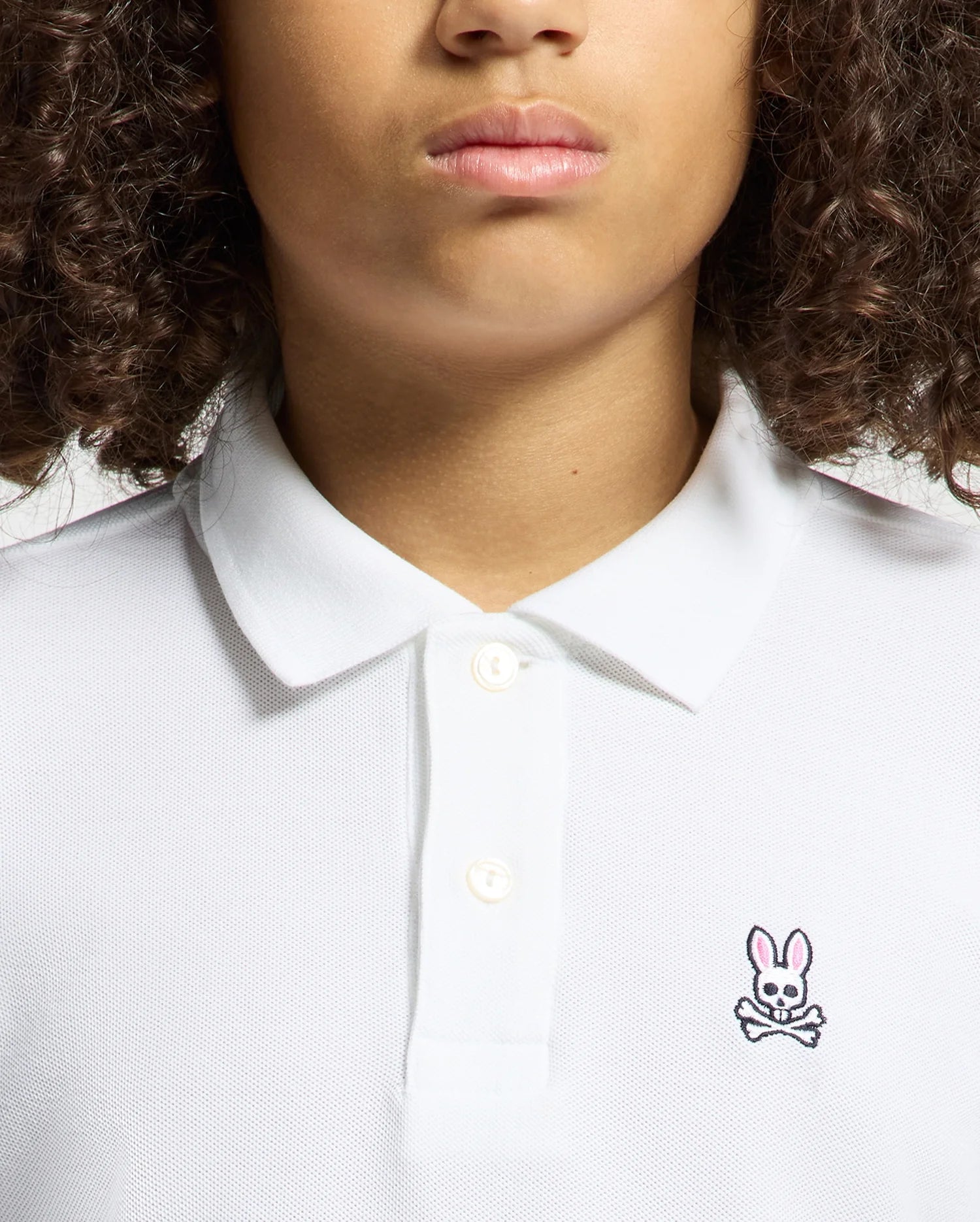 A curly-haired person wears a KIDS CLASSIC PIQUE POLO SHIRT by Psycho Bunny, ideal for back-to-school outfits. This white shirt stands out with its unique chest logo—a pink-eared rabbit above crossed bones.