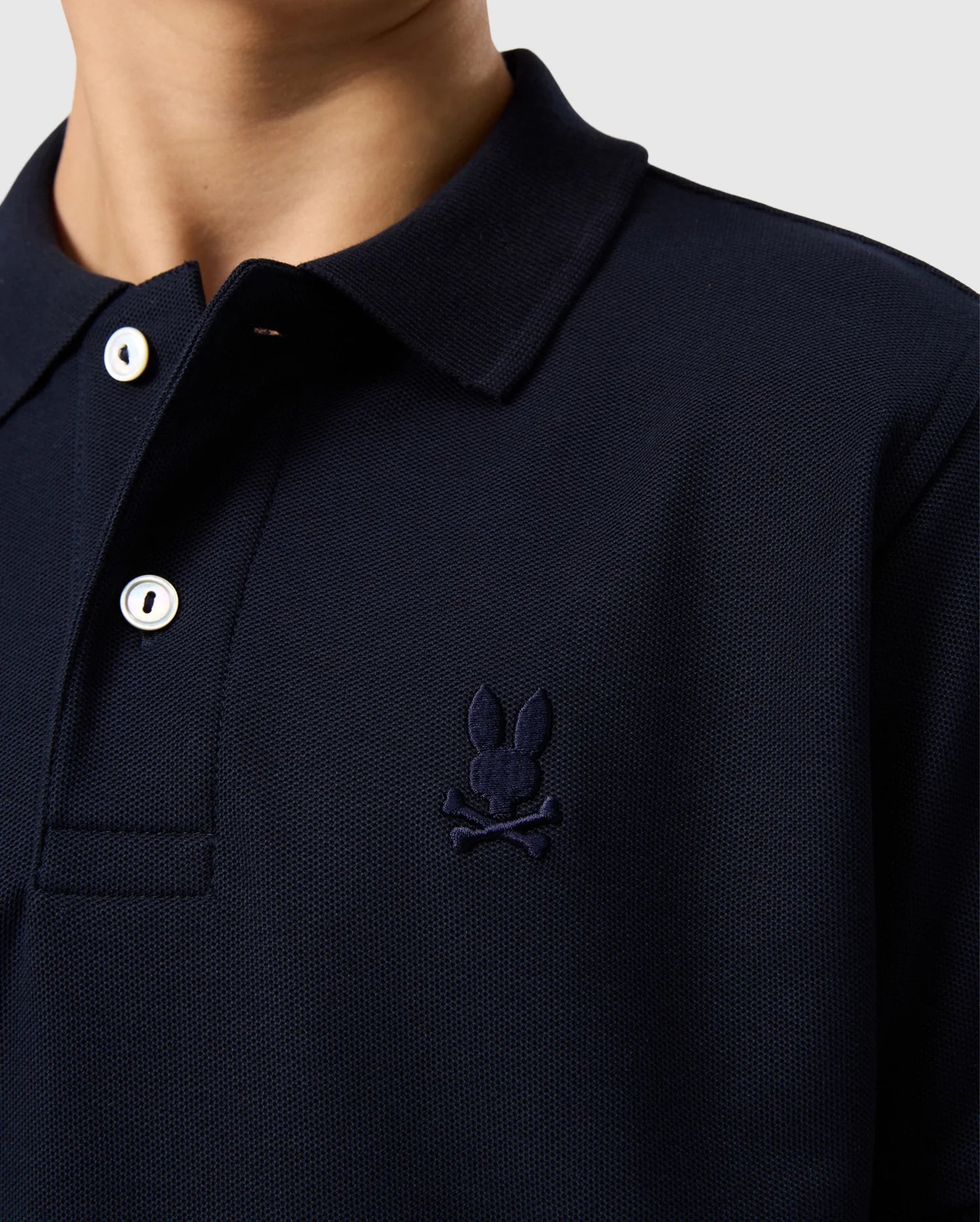 Close-up image of a person wearing the KIDS CLASSIC PIQUE POLO SHIRT by Psycho Bunny in navy. This polo is crafted from soft Pima cotton and features an embroidered Bunny and crossbones emblem on the left chest, complemented by white buttons.