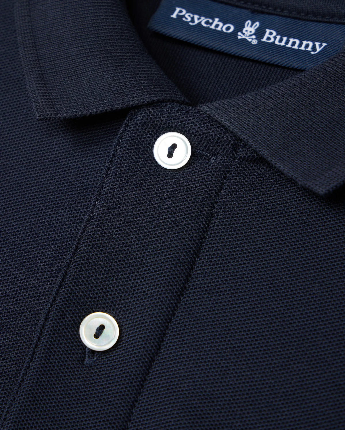 This image shows a detailed view of the Kids Classic Pique Polo Shirt in dark blue, made from Pima cotton. The design includes an embroidered Bunny logo on the chest and a label with "Psycho Bunny" branding. The shirt is accented by two white buttons on the front placket.