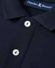 This image shows a detailed view of the Kids Classic Pique Polo Shirt in dark blue, made from Pima cotton. The design includes an embroidered Bunny logo on the chest and a label with "Psycho Bunny" branding. The shirt is accented by two white buttons on the front placket.