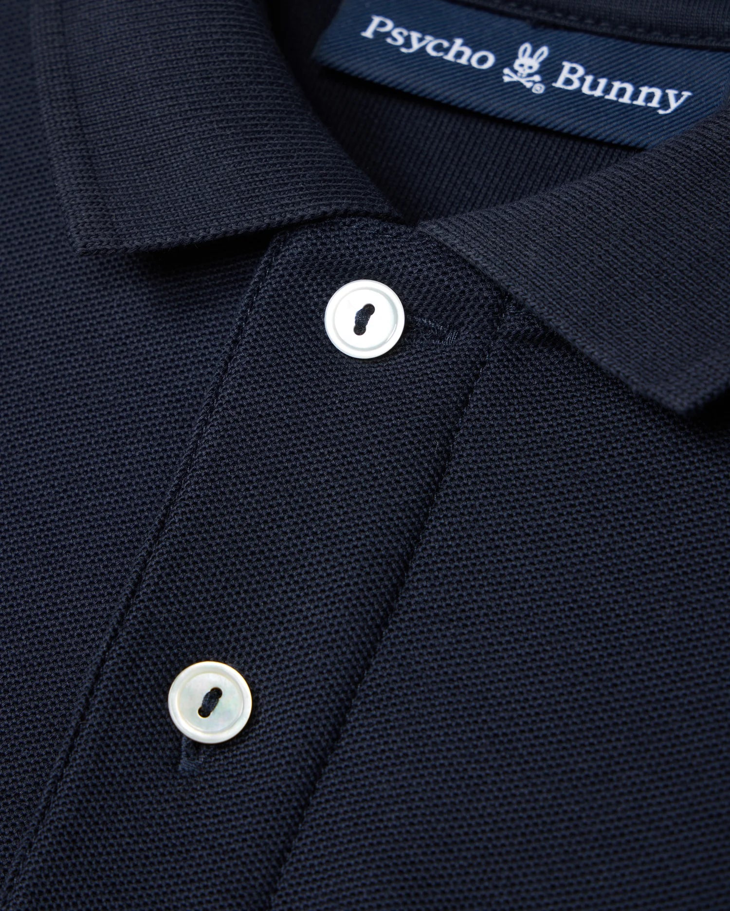 This image shows a detailed view of the Kids Classic Pique Polo Shirt in dark blue, made from Pima cotton. The design includes an embroidered Bunny logo on the chest and a label with "Psycho Bunny" branding. The shirt is accented by two white buttons on the front placket.