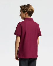 A young boy stands facing away, dressed in a stylish Psycho Bunny Kids Brandson Pique Polo in burgundy paired with dark pants. His hair is short and styled. The plain and neutral background accentuates the classic look of his soft Pima cotton polo.