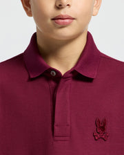 A person dons the Psycho Bunny KIDS BRANDSON PIQUE POLO in maroon, crafted from soft Pima cotton with a buttoned collar. This stylish polo shirt for kids features an embroidered rabbit and crossbones logo on the chest, contrasting against its plain, light-colored background.