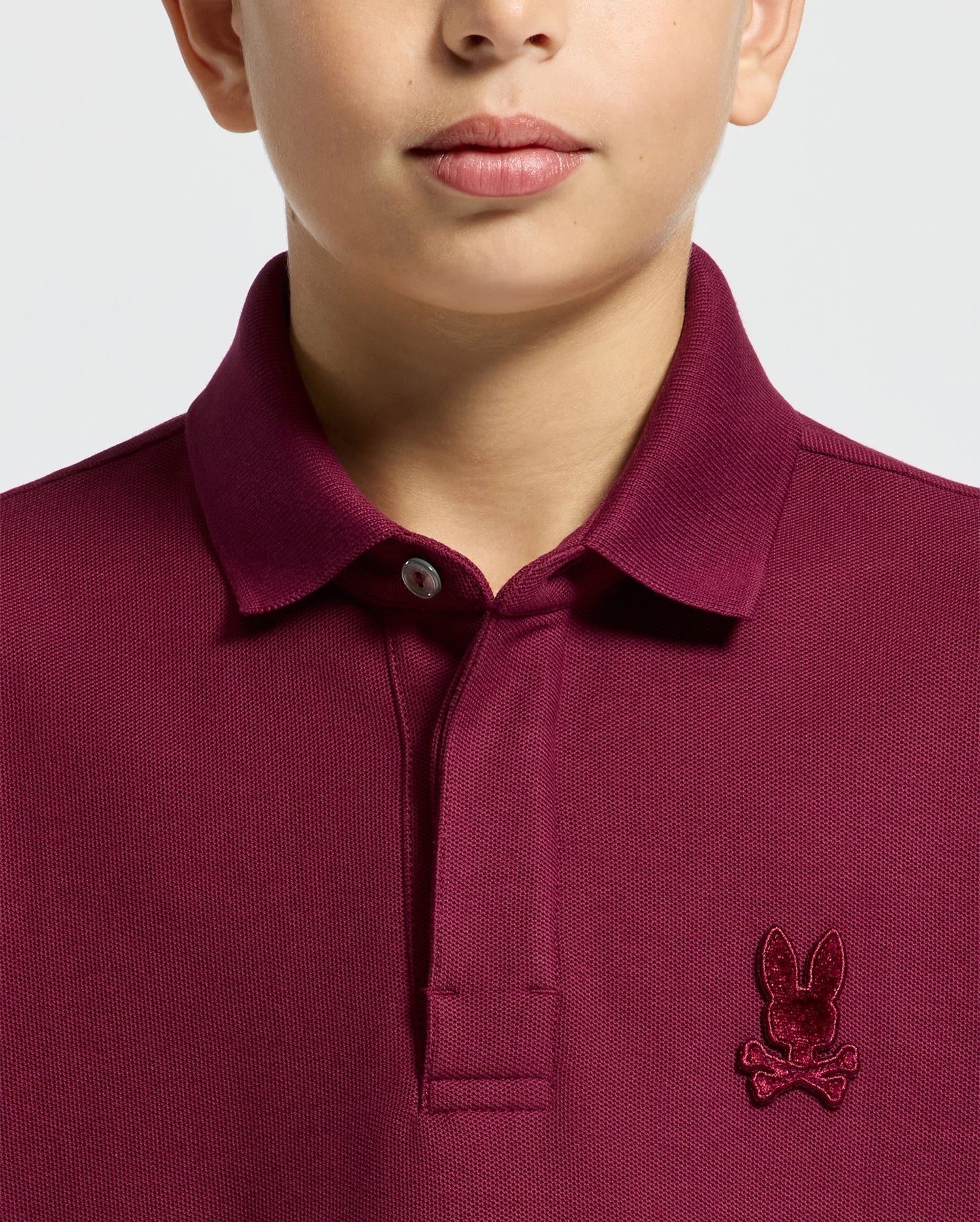 A person dons the Psycho Bunny KIDS BRANDSON PIQUE POLO in maroon, crafted from soft Pima cotton with a buttoned collar. This stylish polo shirt for kids features an embroidered rabbit and crossbones logo on the chest, contrasting against its plain, light-colored background.