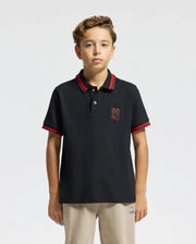 A young person with short curly hair is wearing the Psycho Bunny KIDS PIERCE PIQUE POLO - B0K201E200, which features premium quality material and red stripes on the collar and sleeves. This Pima cotton polo is adorned with an embroidered chest bunny logo. They are standing against a plain white background.