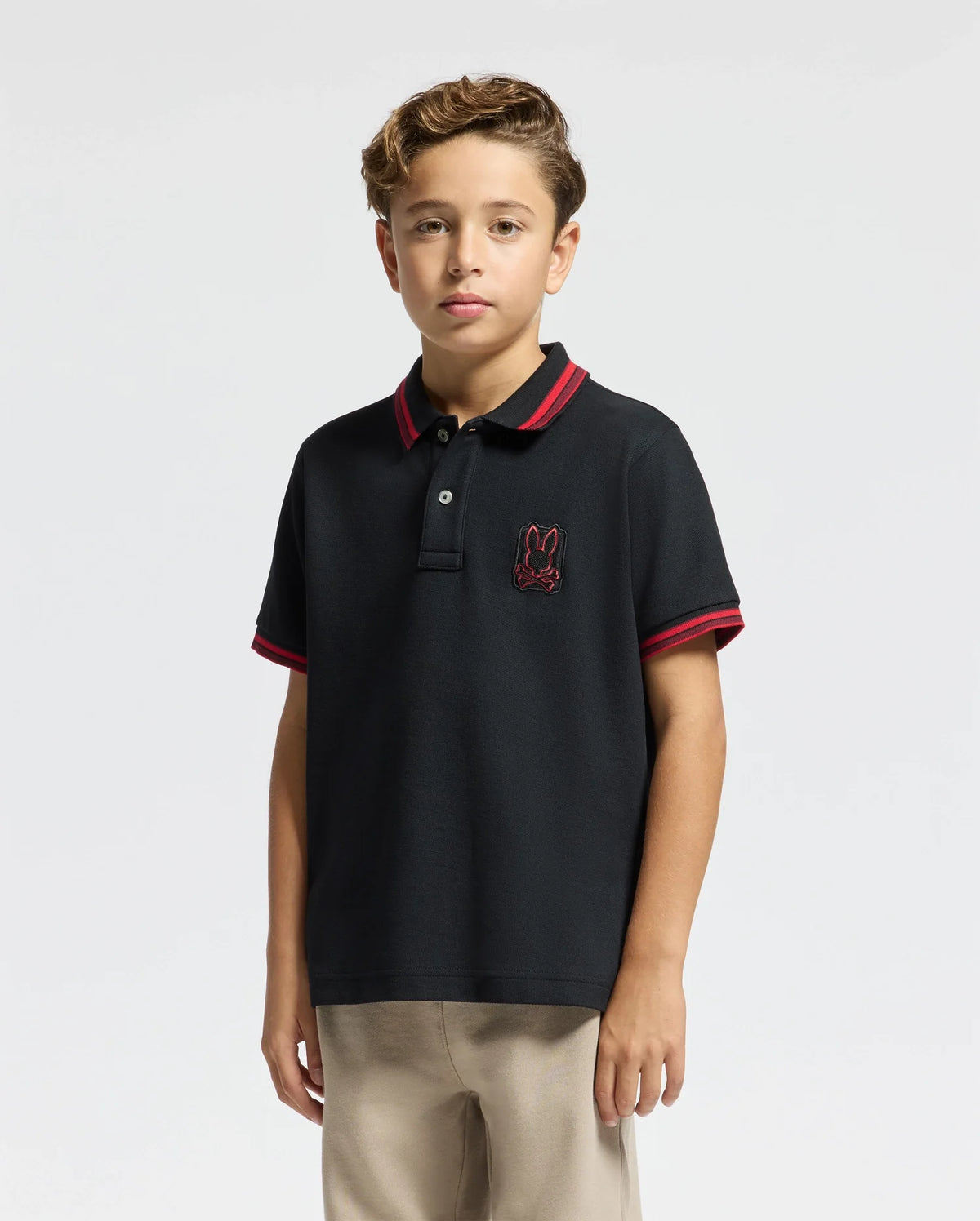 A young person stands against a plain background, wearing the KIDS PIERCE PIQUE POLO - B0K201E200 by Psycho Bunny. The premium quality black polo shirt features a red trim and an embroidered bunny on the chest. With its charming design, this Pima cotton polo complements their confident look as their short, wavy hair frames their face.