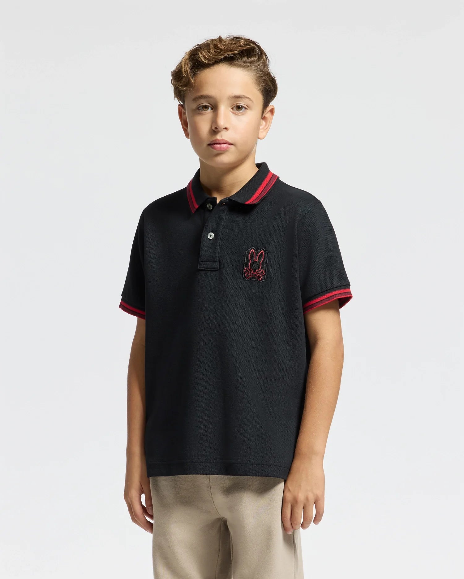 A young person stands against a plain background, wearing the KIDS PIERCE PIQUE POLO - B0K201E200 by Psycho Bunny. The premium quality black polo shirt features a red trim and an embroidered bunny on the chest. With its charming design, this Pima cotton polo complements their confident look as their short, wavy hair frames their face.