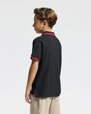 A young boy with short hair stands sideways, wearing the KIDS PIERCE PIQUE POLO - B0K201E200 by Psycho Bunny, a black polo shirt with red trim and beige pants. This premium quality polo features an embroidered chest bunny logo, adding a playful touch against the plain white background.