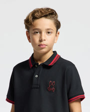 A young boy with curly hair stands facing the camera, wearing the KIDS PIERCE PIQUE POLO - B0K201E200. The black shirt from Psycho Bunny features red trim and an embroidered chest bunny logo. The background is plain white.