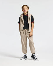 A young person stands wearing a black Psycho Bunny KIDS PIERCE PIQUE POLO with red trim, beige pants, and a matching beige sweater draped over their shoulders. They have short curly hair and are sporting black high-top sneakers. The background is plain white.