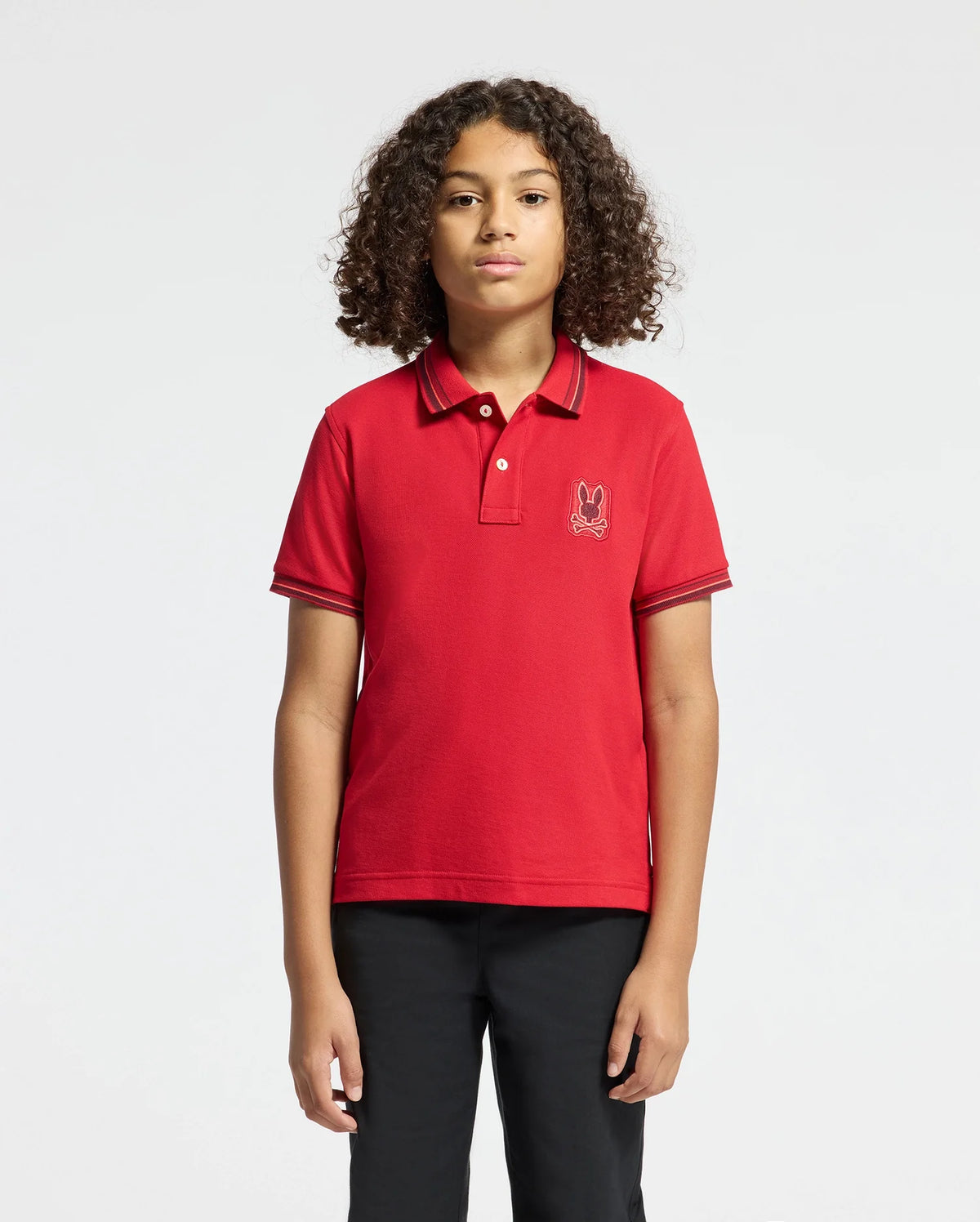 A young person with curly hair stands against a plain background, wearing the KIDS PIERCE PIQUE POLO - B0K201E200 from Psycho Bunny. This stylish red polo, crafted from rich Pima cotton, features an embroidered chest Bunny emblem and is perfectly paired with sleek black pants.